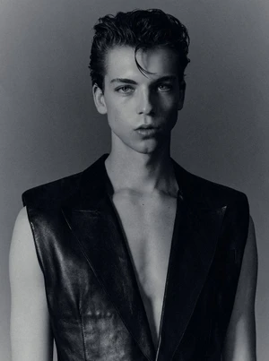 Portrait of a young male model with styled dark hair wearing a black vest against a plain grey studio backdrop.