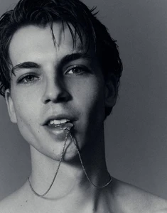 Close-up portrait of a young man biting a silver necklace chain against a neutral studio background