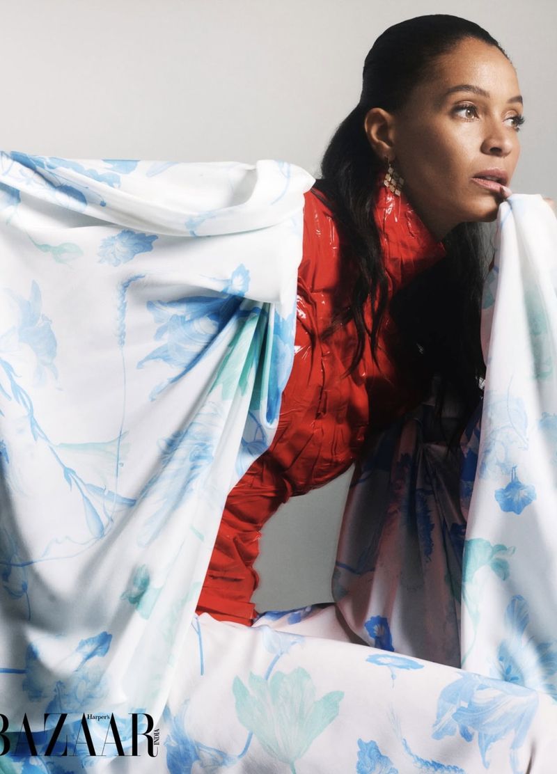 Woman in red top poses behind floral fabric in a studio. Fashion editorial shot with soft lighting showcases clothing and model.
