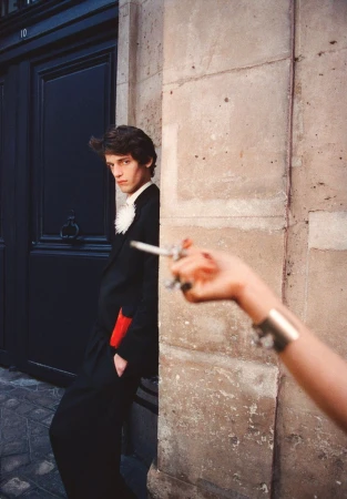 Man in a black suit standing near a dark door with another hand holding a cigarette in the foreground