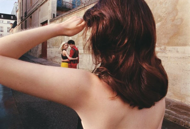 Close-up of a woman's shoulder and dark hair with a couple embracing on a street in the background