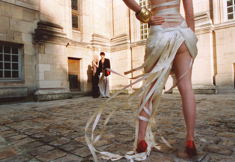 Model in a draped white dress with trailing ribbons and red heels stands in a stone courtyard with figures in the background.