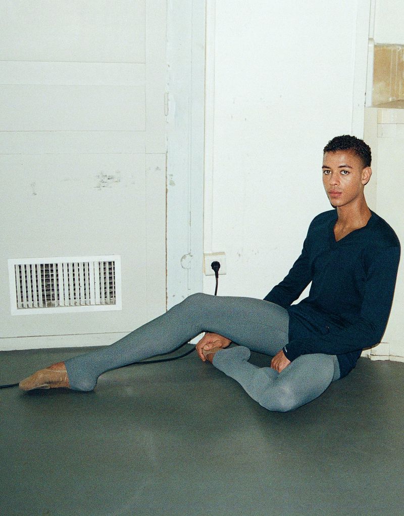 Ballet dancer Guillaume Diop seated on a dark floor wearing a dark blue long-sleeve top and grey tights against a white wall.
