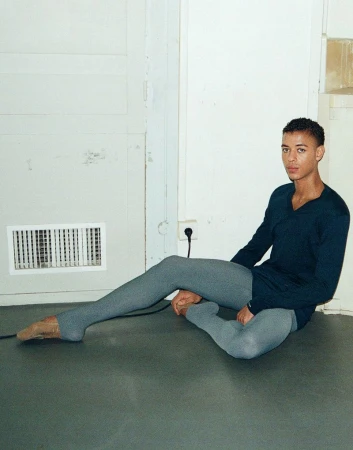 Ballet dancer Guillaume Diop seated on a dark floor wearing a dark blue long-sleeve top and grey tights against a white wall.