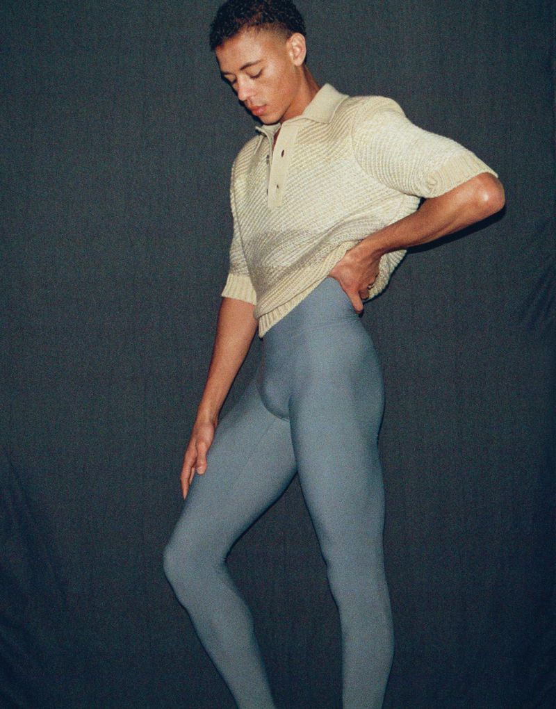 Full-length portrait of dancer Guillaume Diop wearing a cream polo shirt and grey tights against a dark backdrop.
