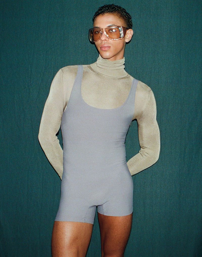 Portrait of Guillaume Diop wearing oversized tinted sunglasses, a turtleneck, and a grey romper against a dark green backdrop