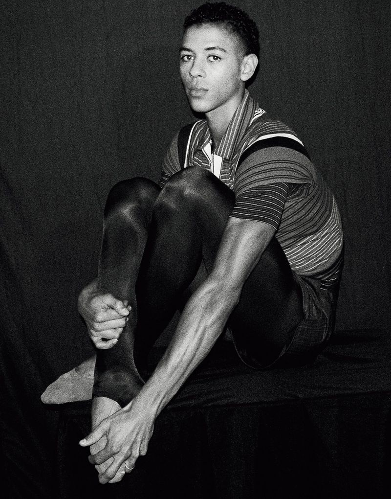 Portrait of Guillaume Diop seated with knees pulled up wearing a striped polo shirt and tights for El Pais Semanal.