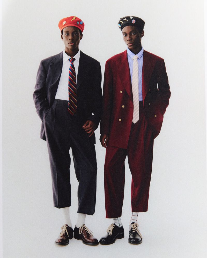 Two dark-skinned men in suits and hats pose in a studio. The man on the left wears a grey suit, while the man on the right is in maroon.