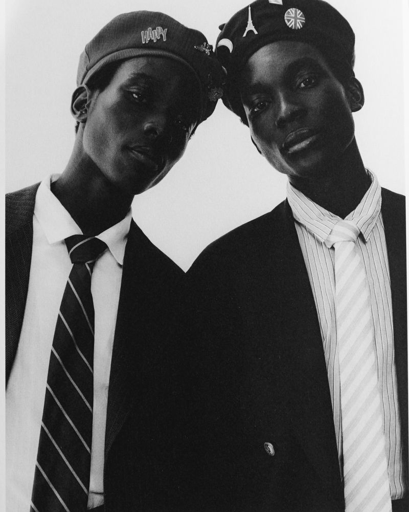 Black and white portrait of two Black models wearing caps, suits, and ties against a plain bright background.