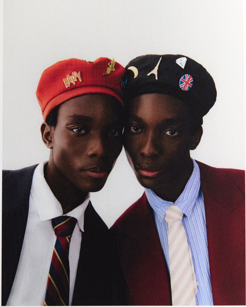 Two male models wearing berets adorned with pins, white shirts, ties, and blazers against a plain background