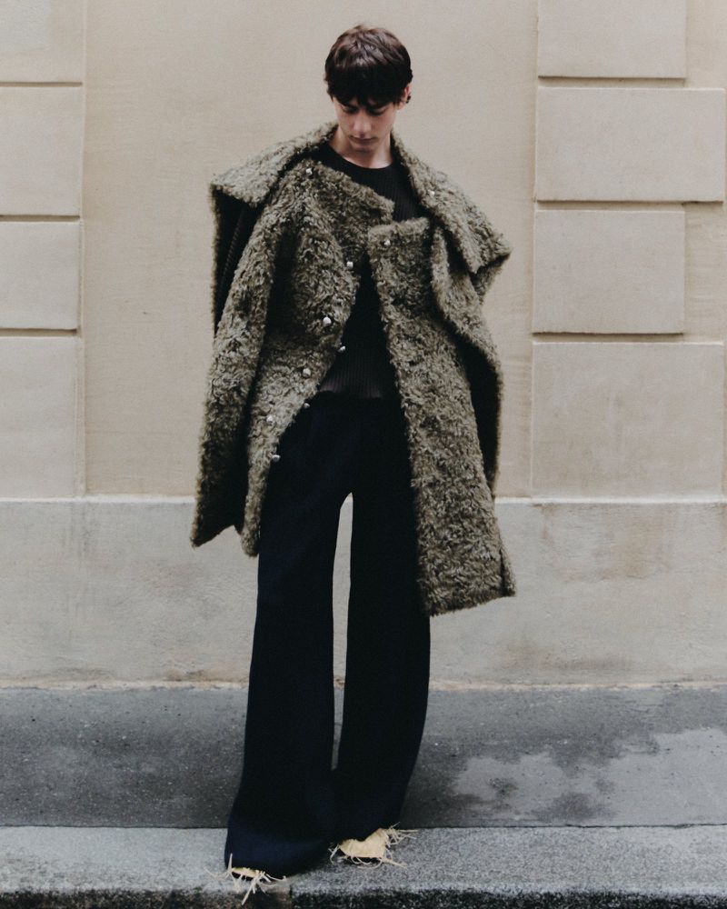 Person in a textured green coat and black pants poses against a neutral wall for a fashion editorial, showcasing textured outerwear.