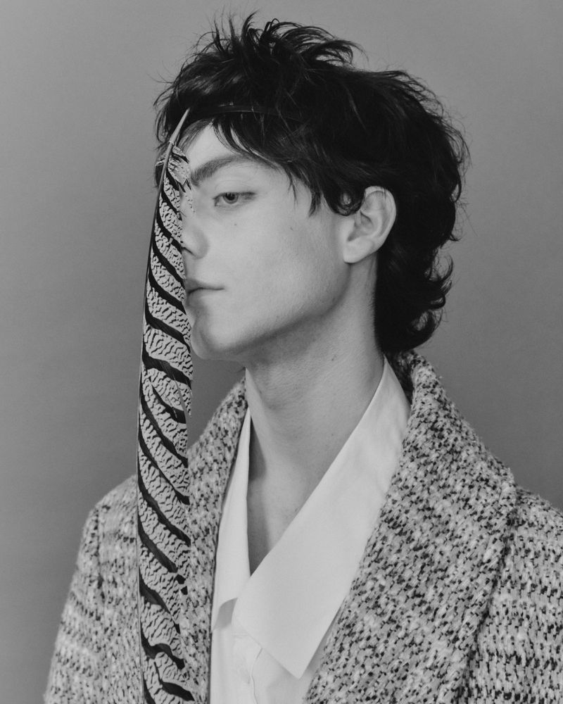Black and white portrait of a young adult male with dark hair, wearing a textured jacket, set against a neutral backdrop with a striped scarf.
