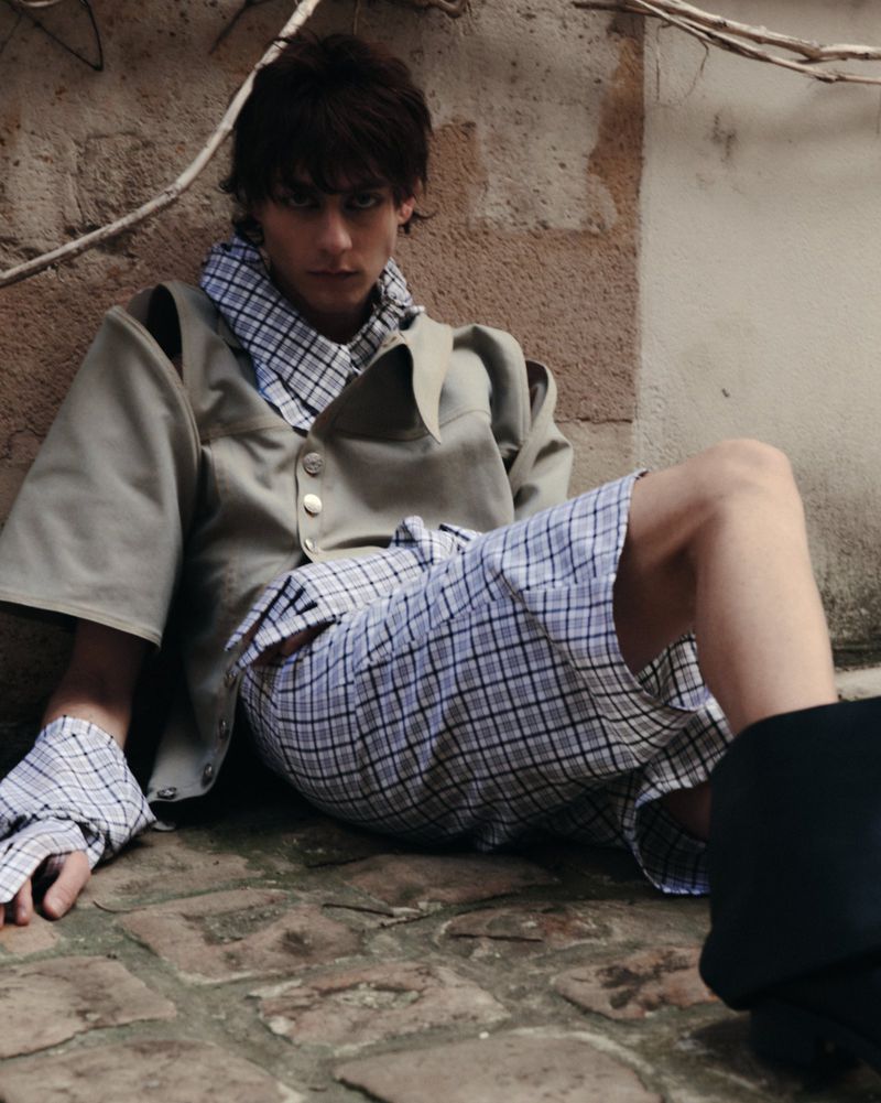 Young adult male in a beige short-sleeve jacket and plaid shorts sits against a textured wall in an outdoor setting, fashion shot.