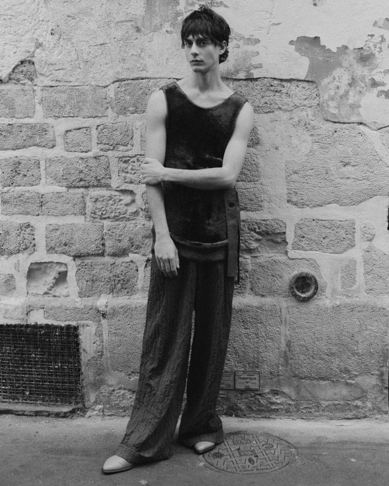 Adult male model posing outdoors in a sleeveless shirt and wide-leg pants, leaning against a stone wall with textured surface.