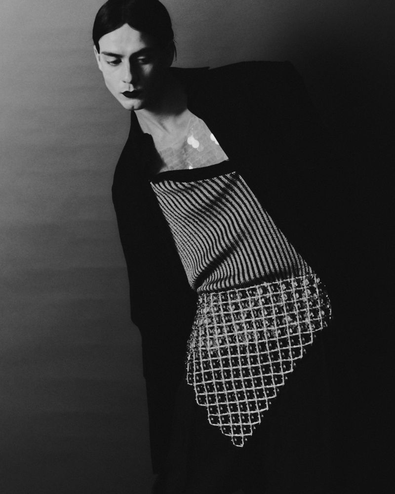 Black and white studio portrait of a person with dark makeup wearing a striped shirt with a beaded overlay and a black jacket.