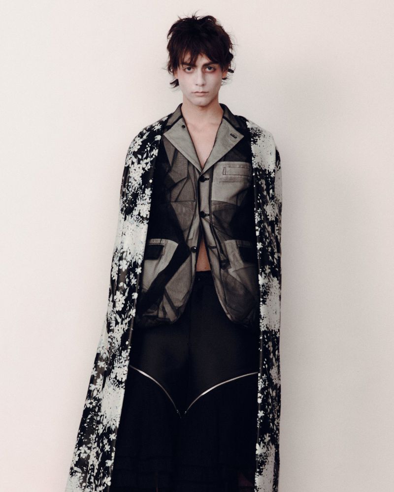 A person with dark hair and dramatic makeup poses indoors wearing a patterned top and long coat against a plain backdrop.