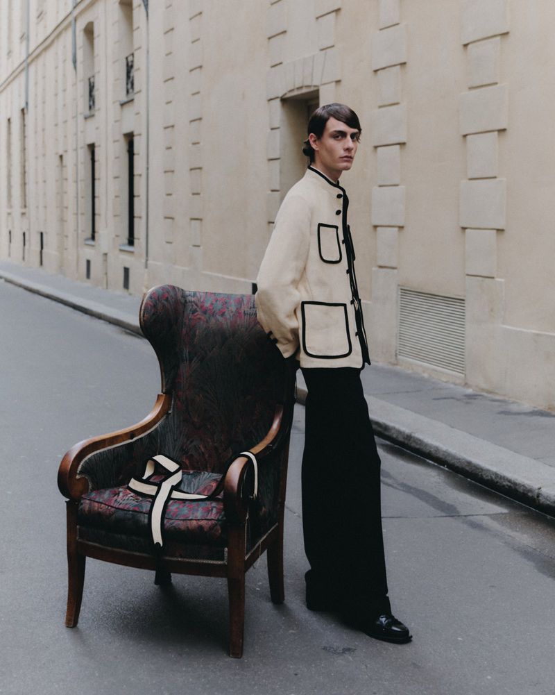A slender man in a cream jacket and black pants poses next to an ornate wingback chair on a European city street with tan buildings.