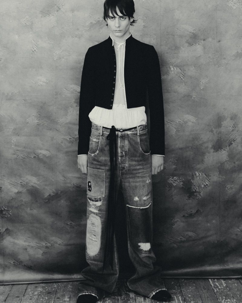 Monochrome portrait of a slender male model wearing a black jacket, sheer white shirt, and distressed denim jeans against a painted backdrop.