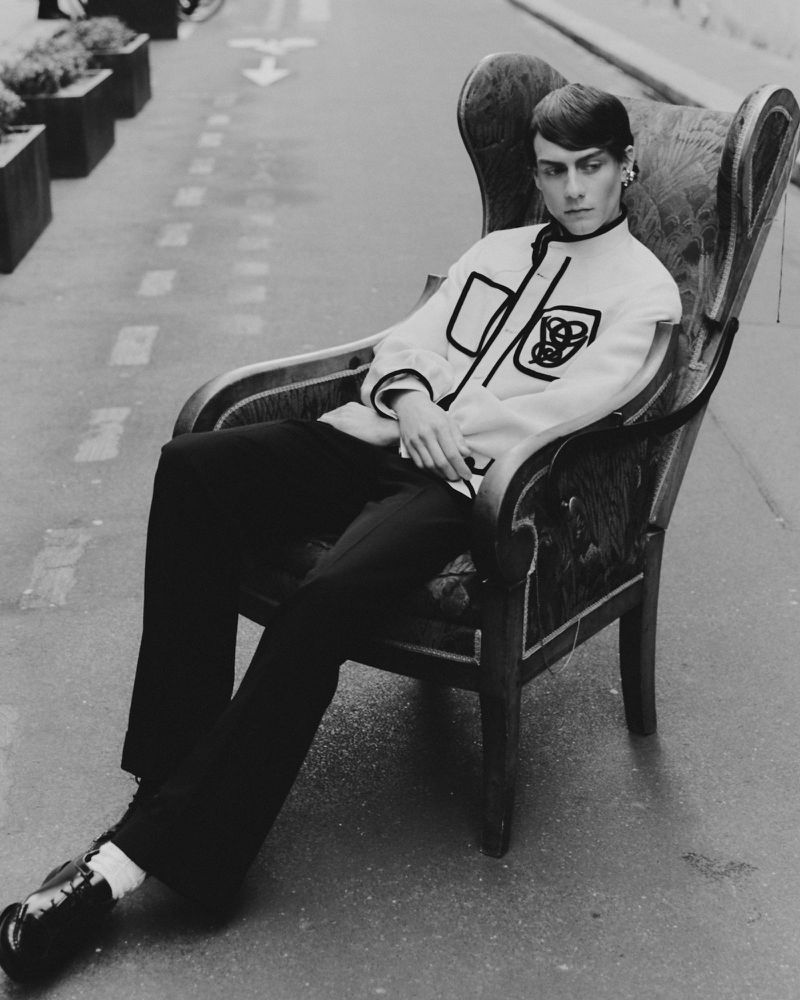 Monochrome shot of a person in a patterned jacket seated in an ornate armchair on a street, creating a striking contrast of textures and styles.