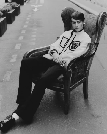 Monochrome shot of a person in a patterned jacket seated in an ornate armchair on a street, creating a striking contrast of textures and styles.