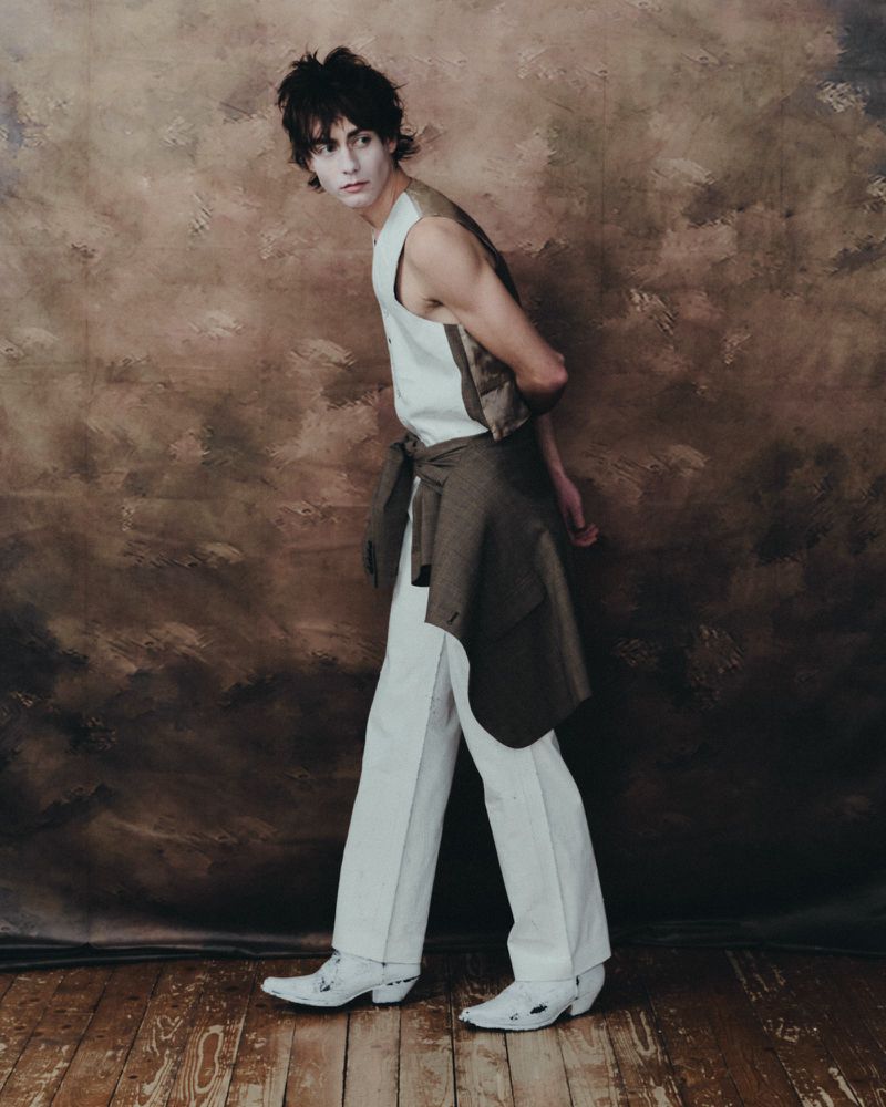 Young adult in vest, white pants, and cowboy boots poses against a textured backdrop on a wood floor in a fashion editorial shot.