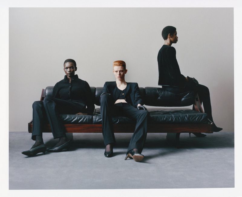 Three models wearing black outfits seated and posing on a dark leather sofa against a neutral grey studio wall.