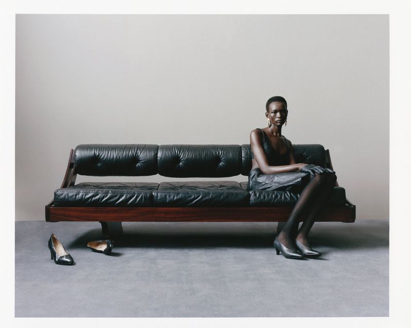 A woman in a black top and skirt sits on a vintage black leather couch, her shoes off to the side, in a fashion editorial shot.
