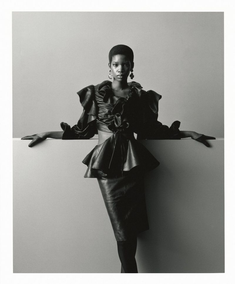 Black and white fashion editorial portrait of a model in a dark ruffled peplum top and skirt posing against a neutral studio wall.
