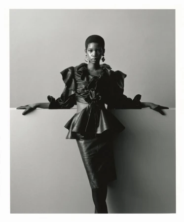 Black and white fashion editorial portrait of a model in a dark ruffled peplum top and skirt posing against a neutral studio wall.