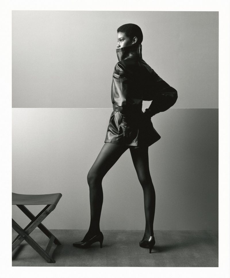 Model in a dark leather jacket and heels posing beside a stool against a split-tone studio background