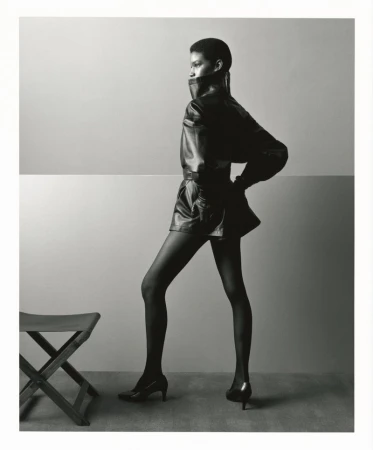 Model in a dark leather jacket and heels posing beside a stool against a split-tone studio background