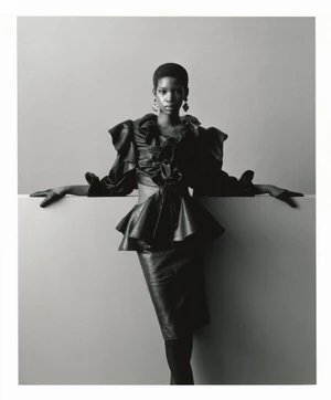 Black and white fashion editorial portrait of a model in a dark ruffled peplum top and skirt posing against a neutral studio wall.