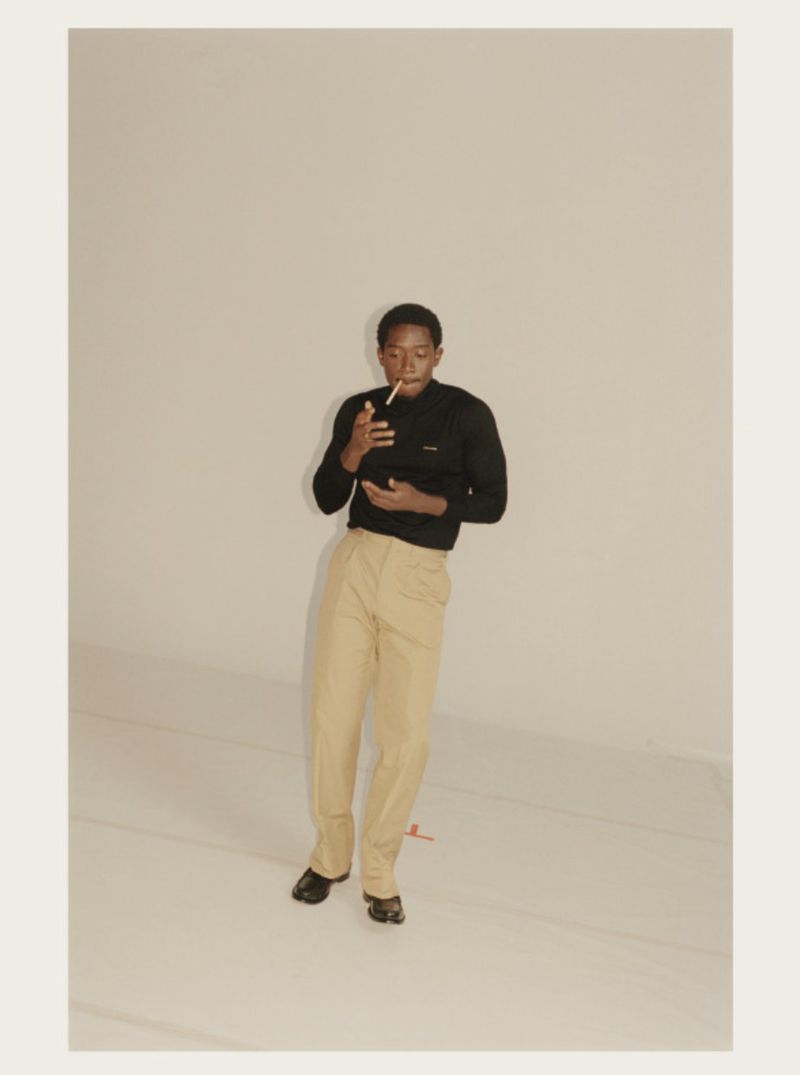 Adult male in a black sweater and khaki pants smoking a cigarette, posed in a photo studio with soft lighting and a neutral background.