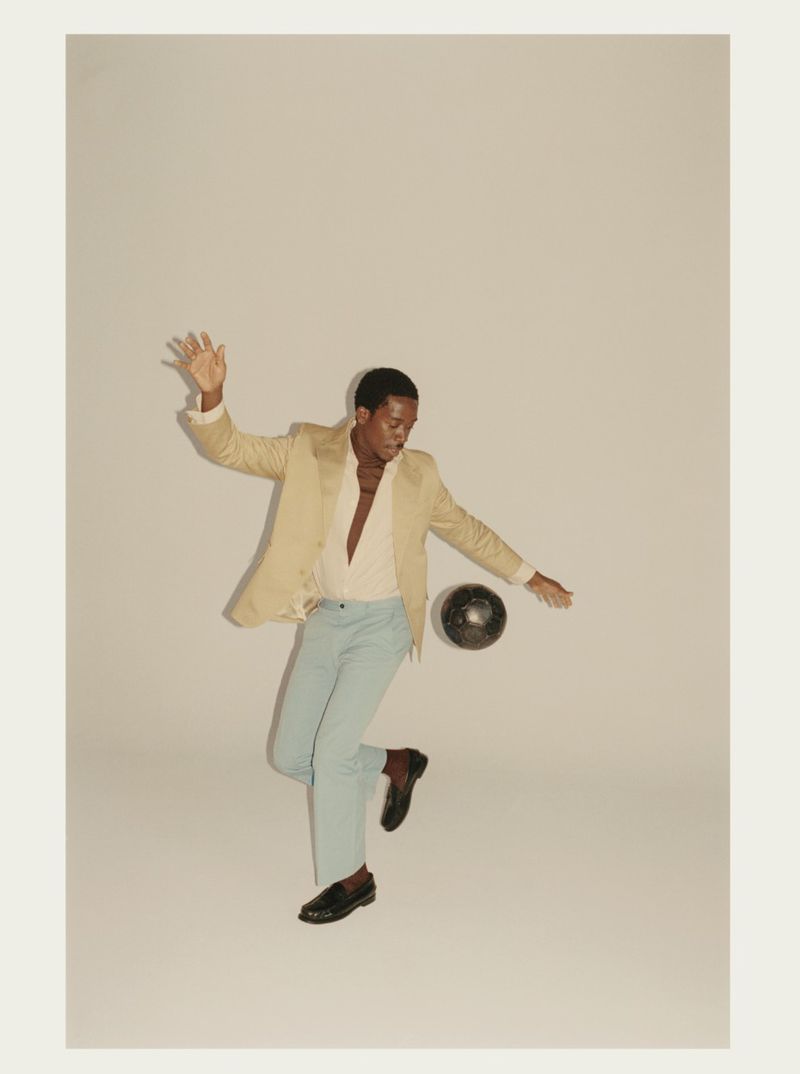 Damson Idris wearing a tan blazer and light blue trousers while interacting with a dark sphere against a neutral background