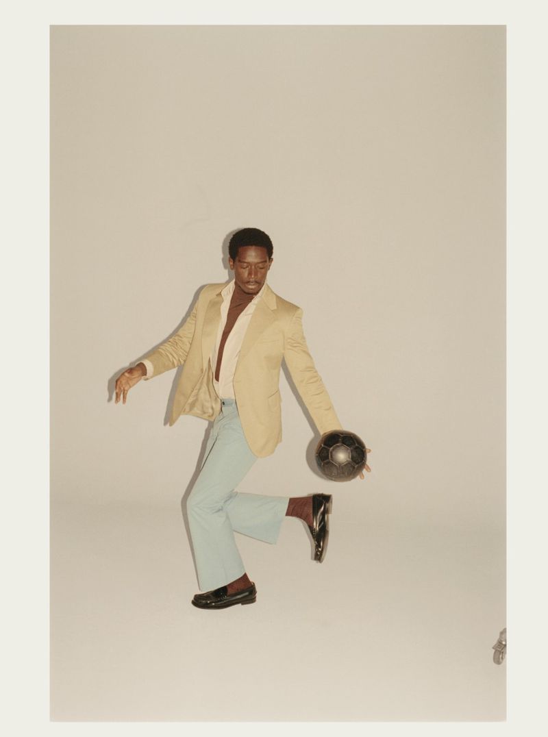 Damson Idris wearing a beige blazer and light blue trousers balancing a soccer ball on his foot against a white background