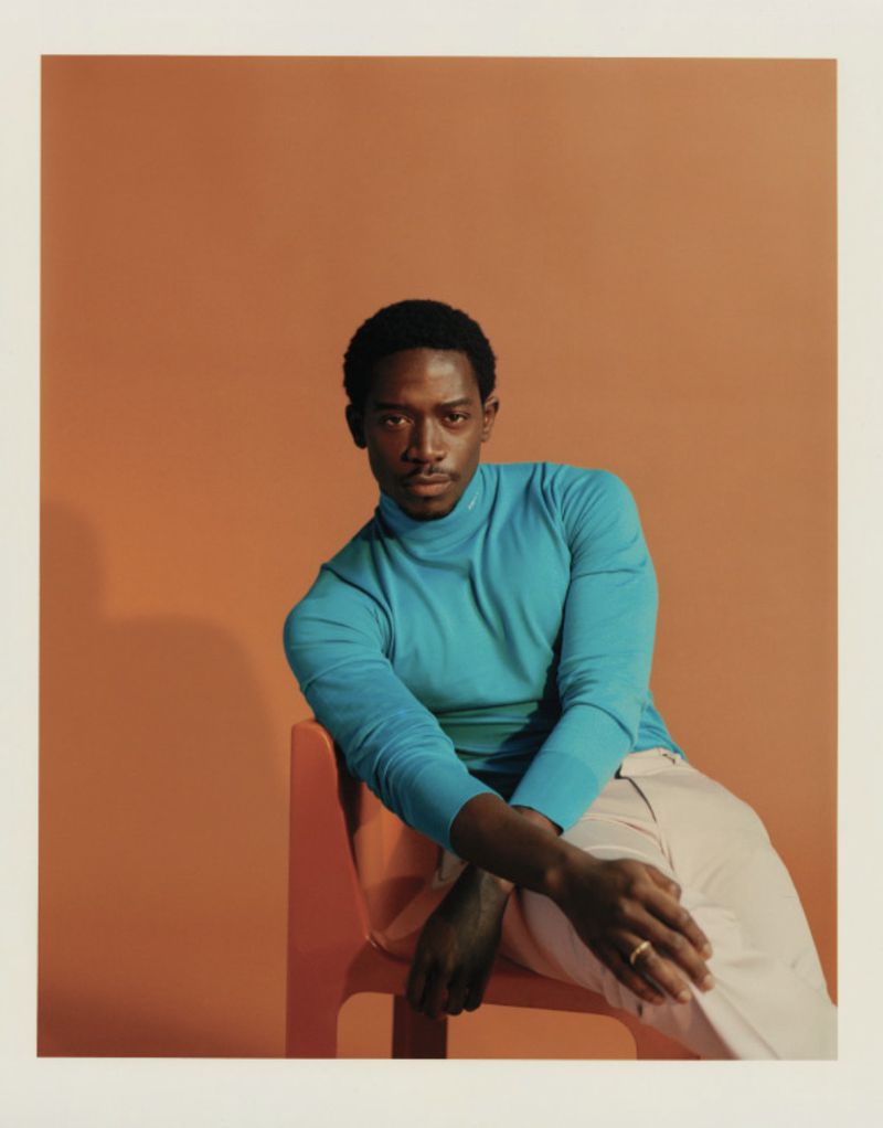 Portrait of Damson Idris wearing a bright blue turtleneck sweater and light trousers seated on an orange chair against a warm brown backdrop.