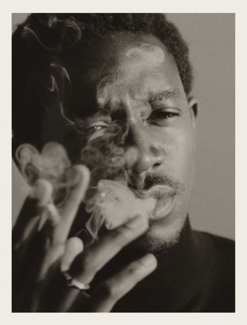 Portrait of Damson Idris exhaling smoke with hand raised holding a cigarette against a plain studio background.