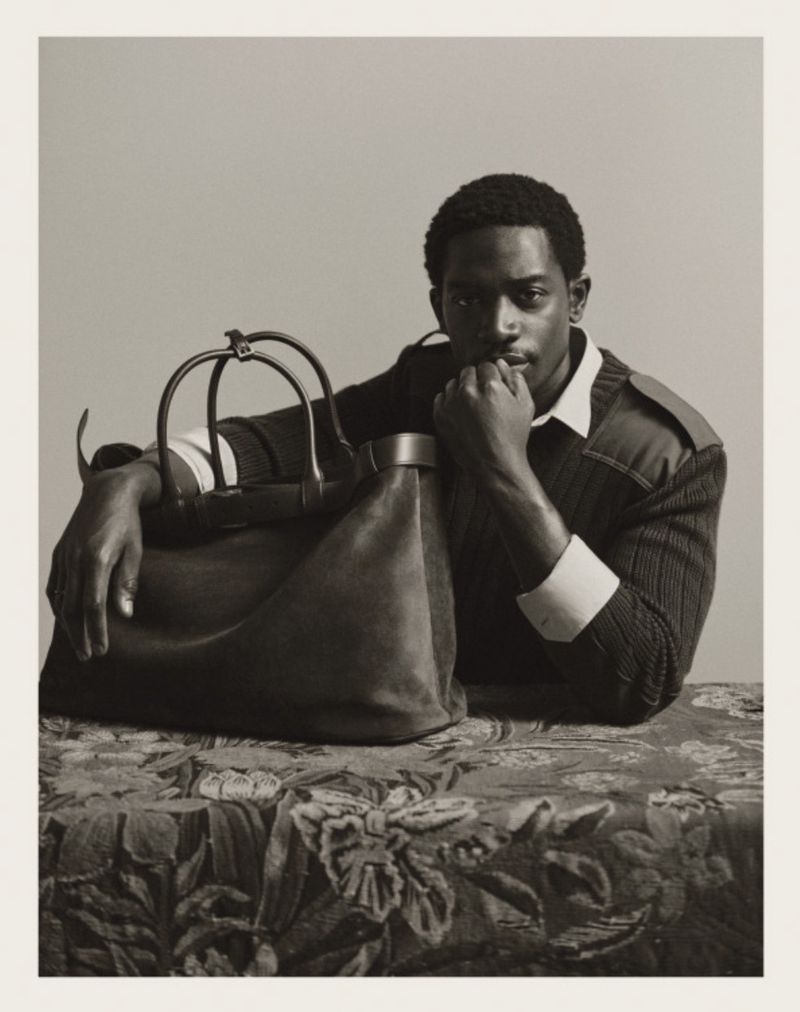 Portrait of Damson Idris resting his chin on his hand beside a large leather holdall bag on a patterned table