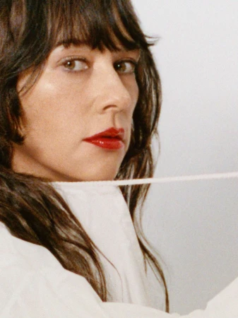 Close-up portrait of Rashida Jones wearing red lipstick and a white top against a seamless white background.