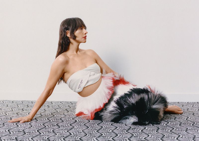 Rashida Jones seated on patterned carpet wearing white strapless top and red-and-white feathered skirt for MARFA Good Friends project