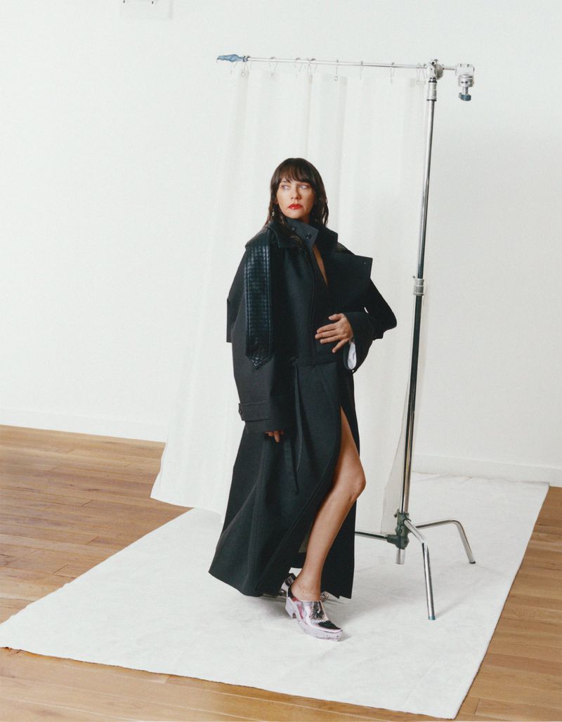 Rashida Jones wearing a long dark coat standing in a studio with a white backdrop and clothing rack for MARFA Good Friends project.