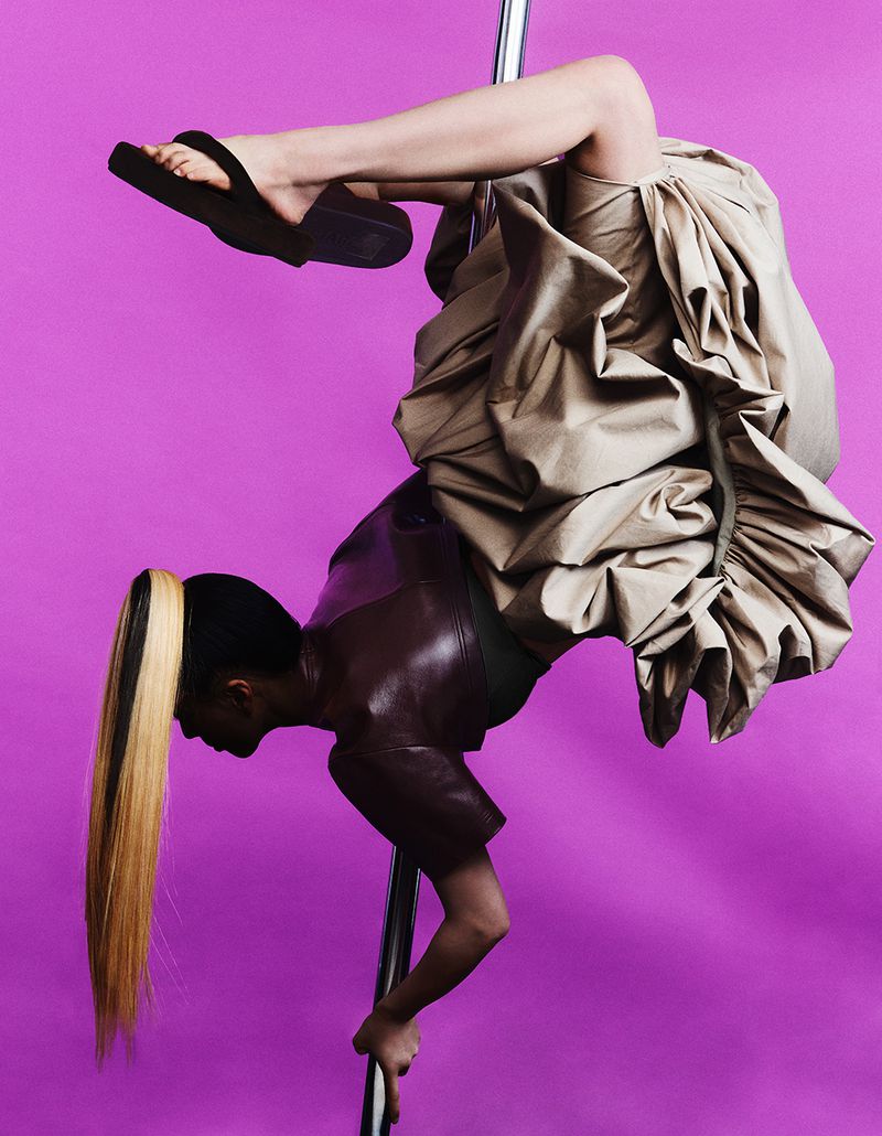 Model performing an upside-down pole dance pose wearing a voluminous skirt and platform sandals against a solid purple backdrop.