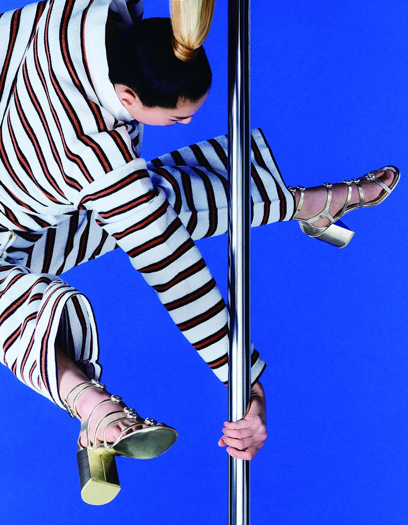 Model in striped clothing and metallic heels climbing a vertical pole against a vibrant blue studio backdrop