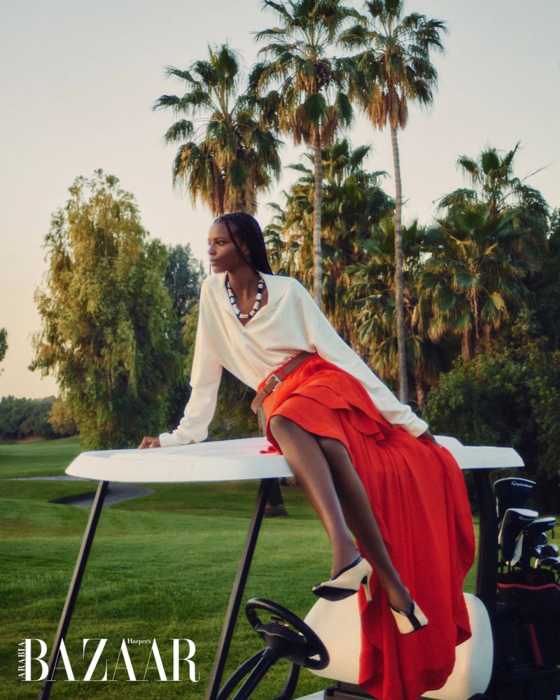 Black woman seated on a golf cart wearing a white blouse and long red skirt outdoors with palm trees.
