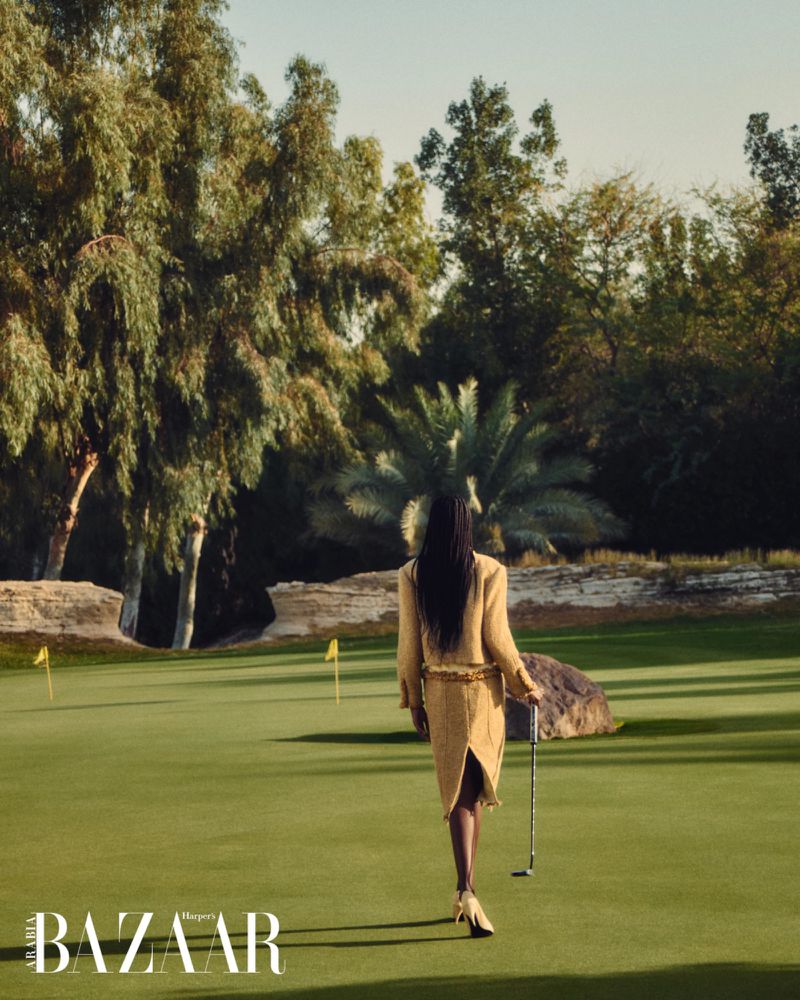 Model wearing a gold textured skirt suit walking away on a green golf course holding a putter, photographed by Vladimir Marti for Harper's Bazaar Arabia