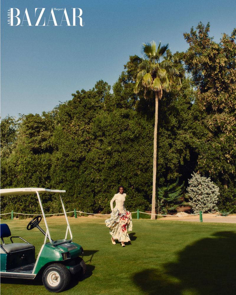 Model in a floral dress walking on a golf course near a green golf cart under a palm tree for Harper's Bazaar Arabia
