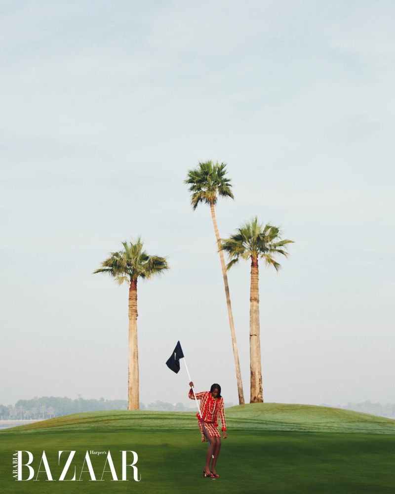Model holding a black golf flag on a green course with three palm trees under an overcast sky for Harper's Bazaar Arabia