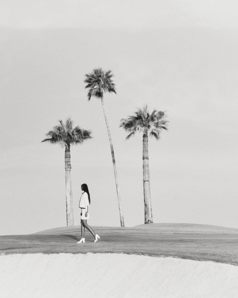 Black and white editorial image of a woman walking past three palm trees on a golf course for Harper's Bazaar Arabia.
