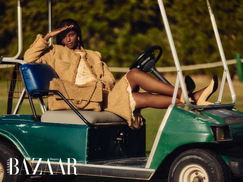 Dark-skinned woman poses in a golf cart wearing a tan Chanel skirt suit and holding her hand up to her forehead for sun protection.