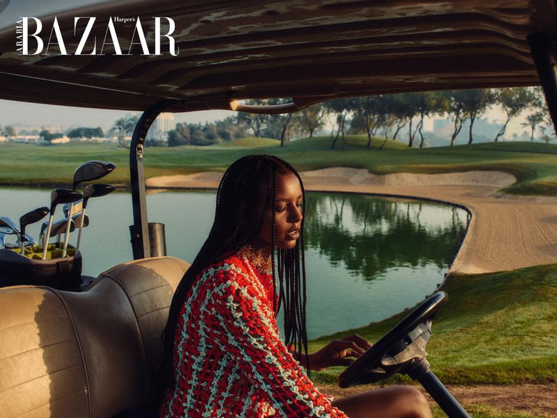 Woman with braided hair wearing a red patterned top seated inside a golf cart overlooking a golf course and water hazard